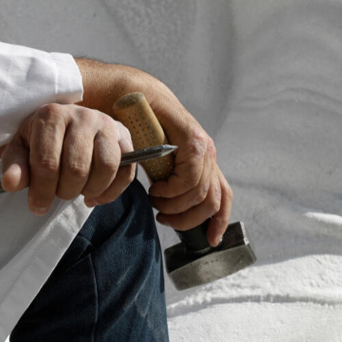 hands of a sculptor wearing white coat and dark blue jeans holding a hammer and chisel resting his arms on his knee with a marble sculpture in the backdrop