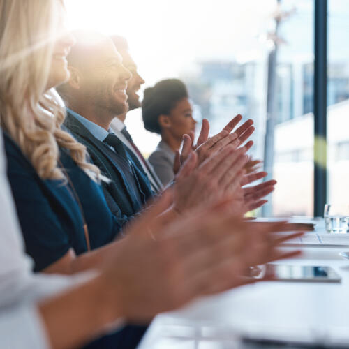 a row of people in business suits with hands clapping and a black woman with an Afro hairstyle deep in the background blurred out